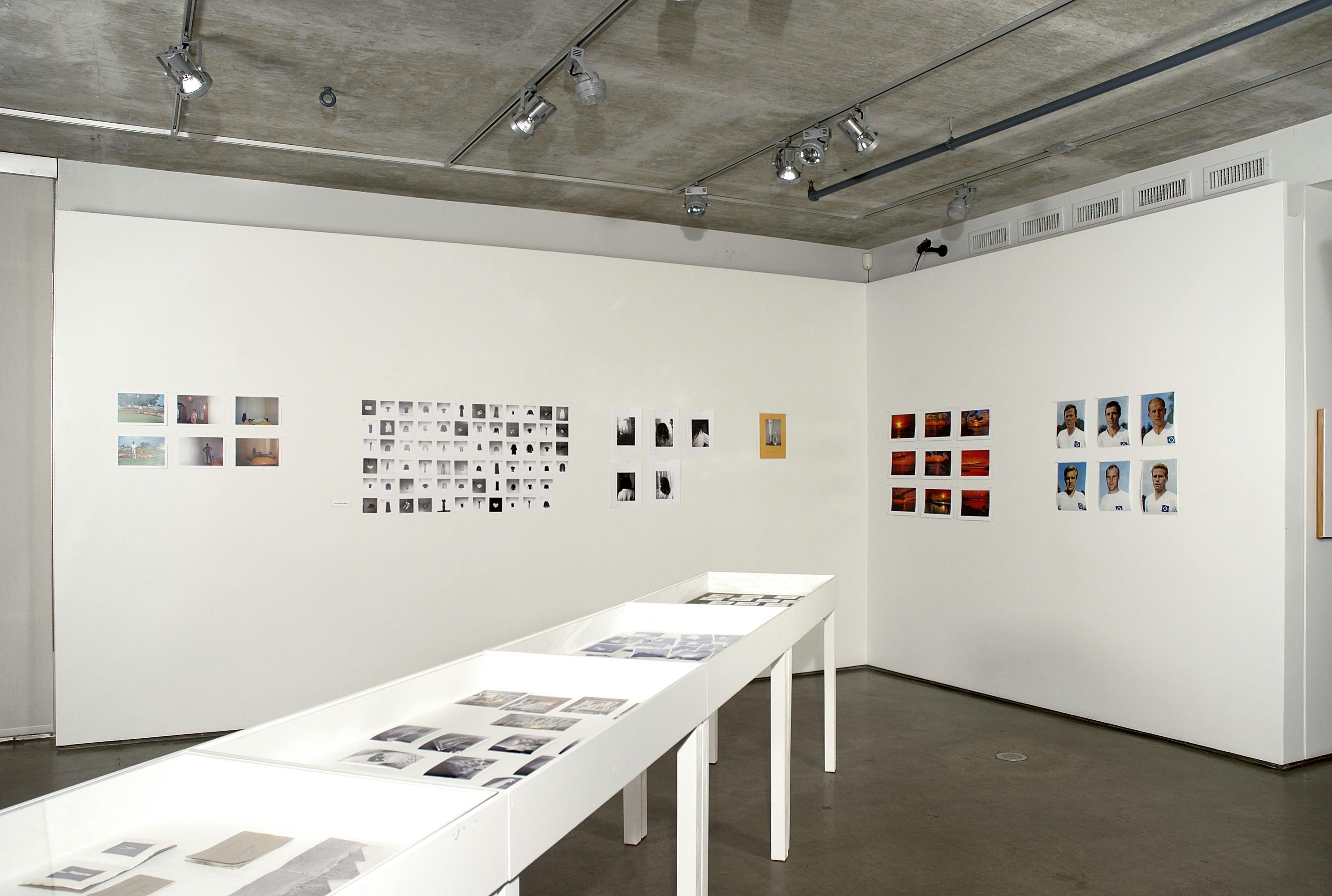 The corner of a gallery, showing dozens of small photos on the walls, divided into three sets. In the space, three long, white vitrine tables are placed together perpendicular to the walls.