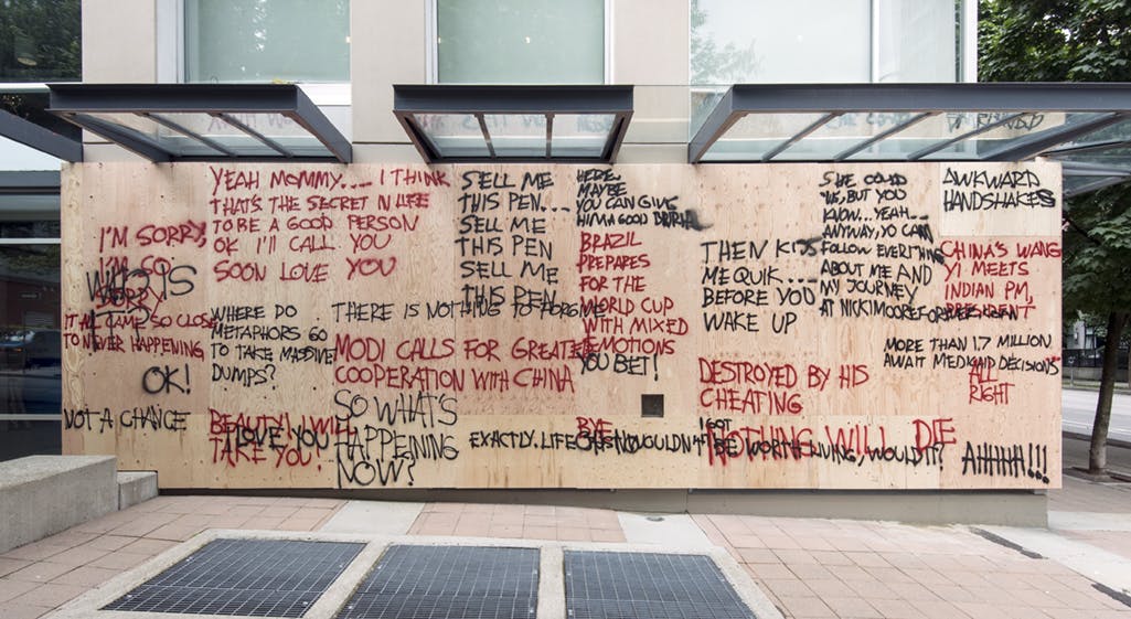 Multiple plywood boards installed on CAG’s facade blocking all the windows. Overlapping spray-painted words in red and black cover the boards. One phrase reads, “AWKWARD HANDSHAKES.” 
