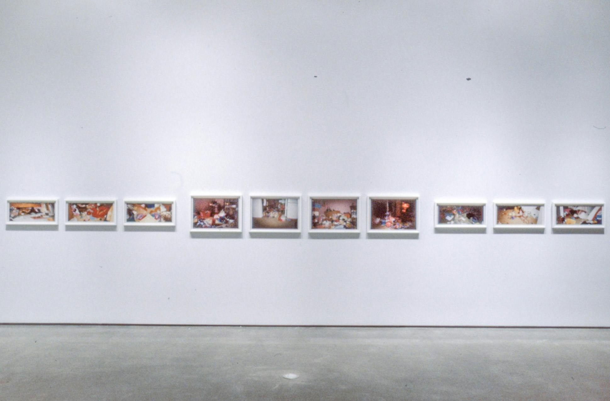 Ten coloured photographs are mounted on the gallery wall. Many of those images depict colourful objects piled up in a room. This room has a white floor and a pink wall.