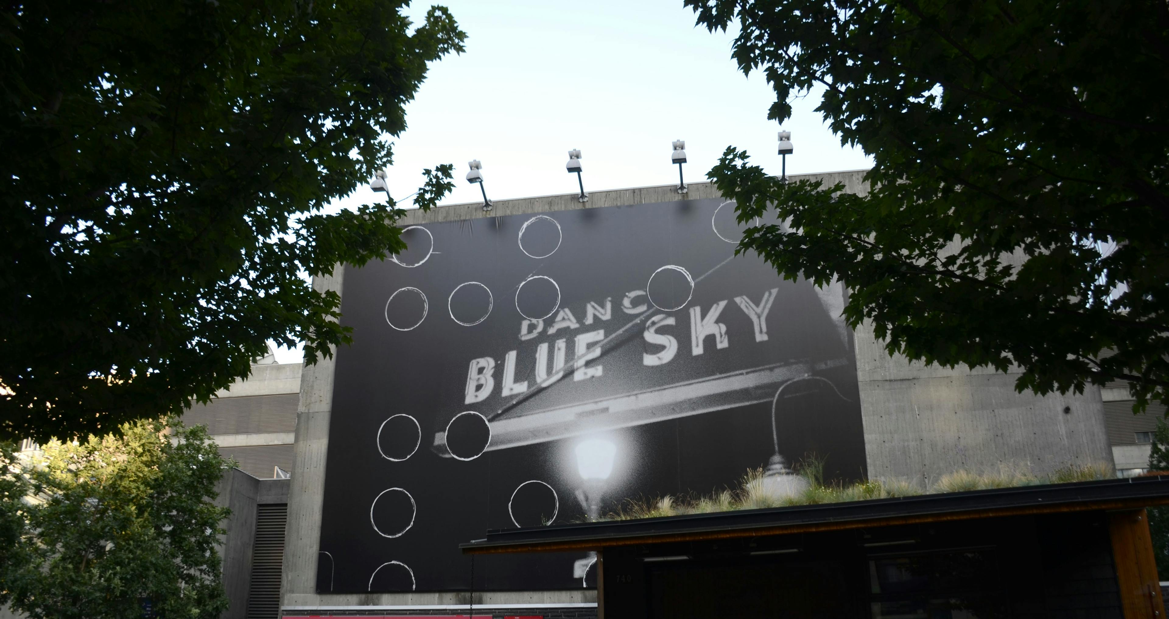 An image of the side of a building framed by two trees in the foreground. Installed on the building is a billboard-sized printed image of a neon sign and street light. The neon sign reads BLUE SKY.
