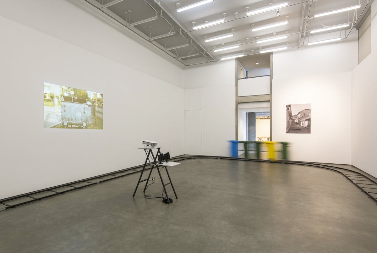 A video projection and a photograph installed on two gallery walls. Five connected garbage bins of various colours move around the gallery on a small rail line installed at the perimeter.