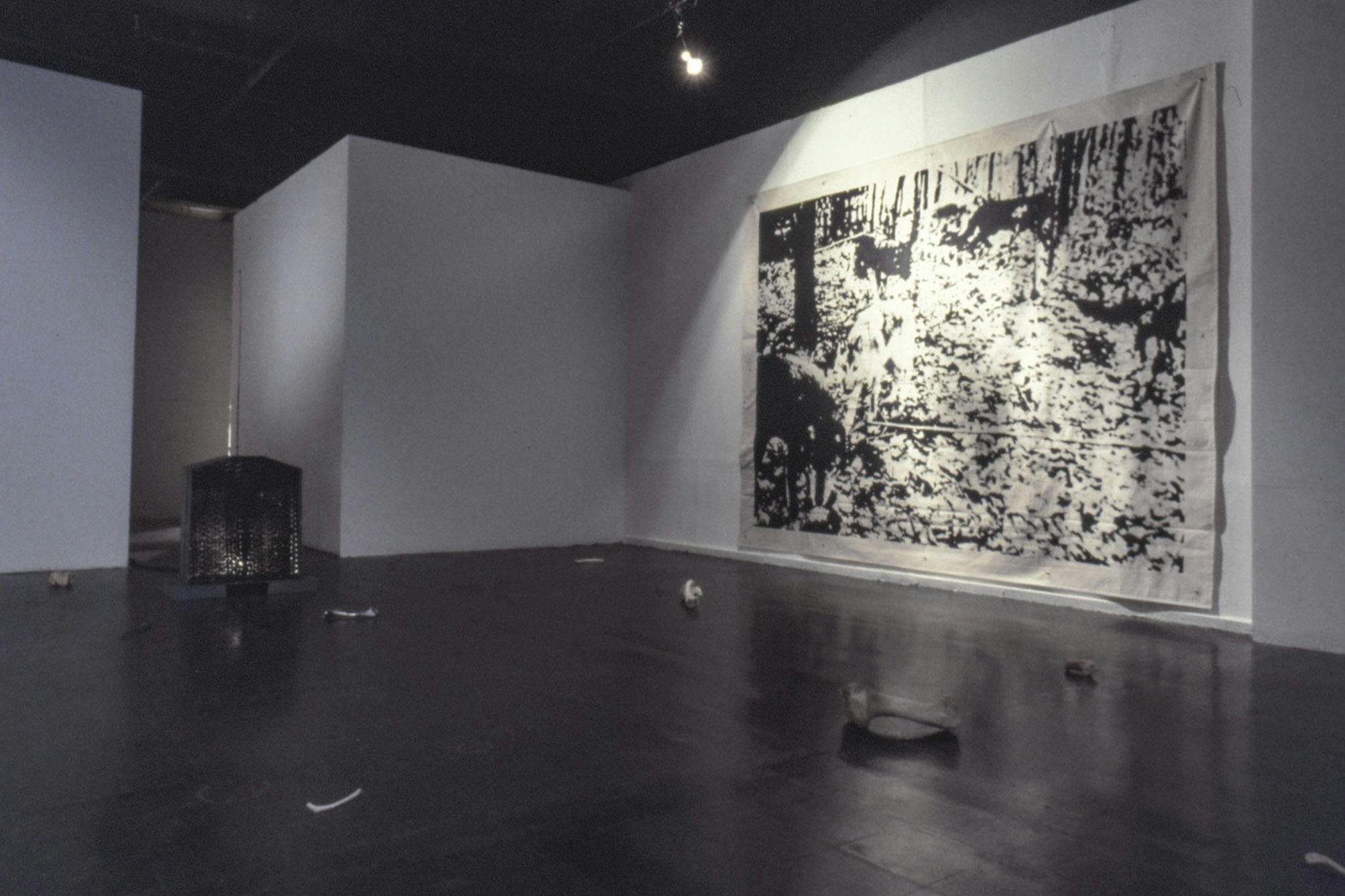On the wall of a dark gallery space, there is a large piece of canvas shows a print of wolves in a forest. On the floor, there is a small glowing speaker as well several natural and cast steel bones.