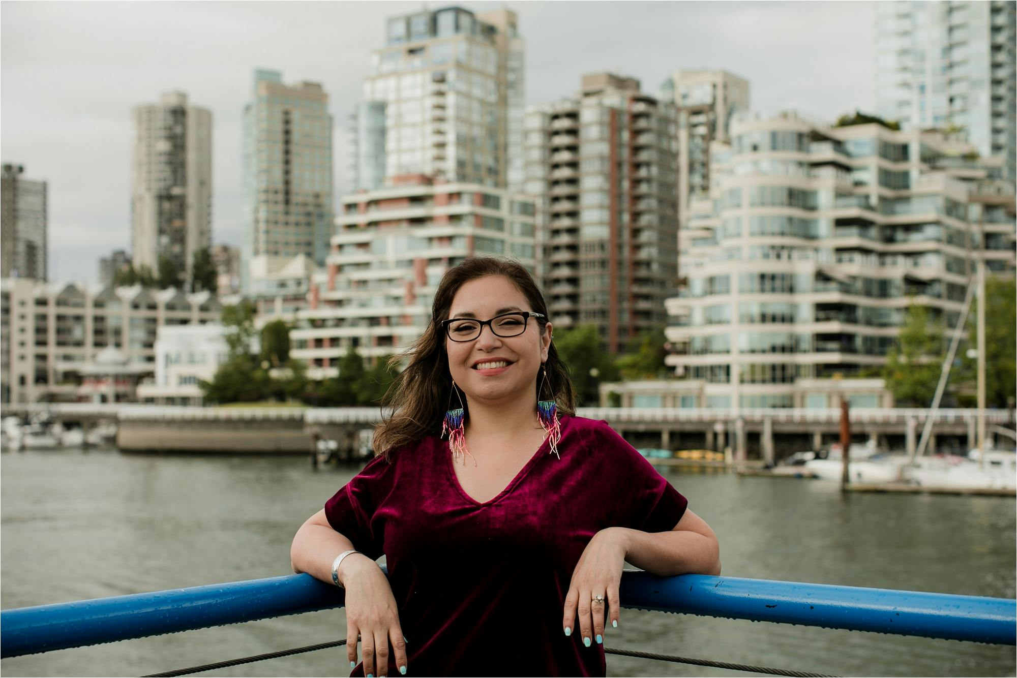 A photograph of Michelle Sound standing and smiling outside by the water. Michelle is wearing a pair of black glasses, beaded earrings, and a wine-red shirt. 
