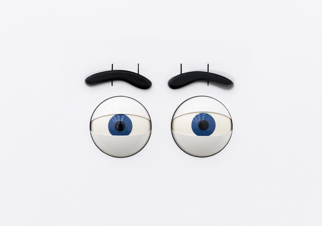 A pair of large, animatronic cartoonish blue eyes with thick black eyebrows and pronounced white eyelids stare out from the image. 