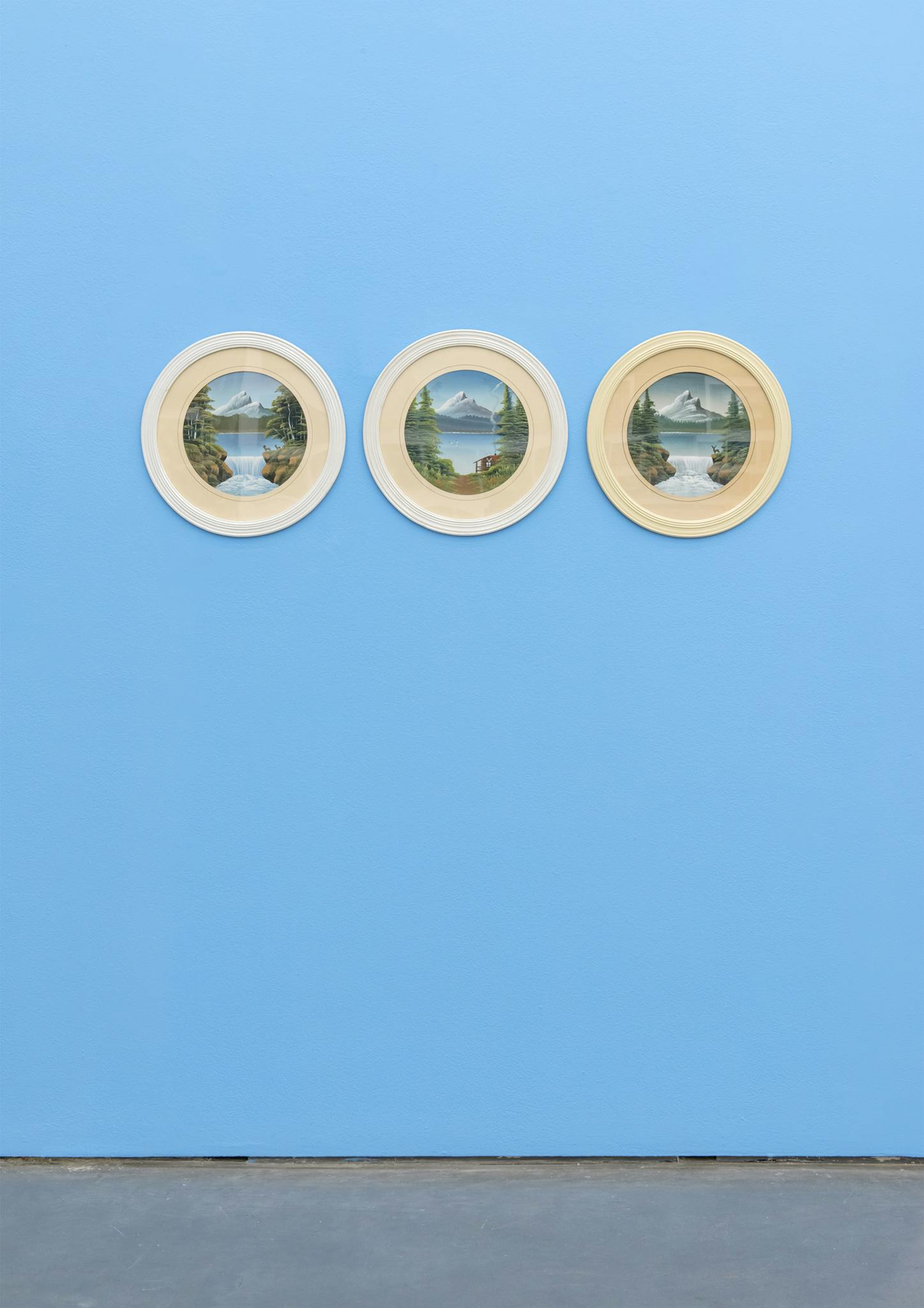 Three circular paintings of mountain landscapes hang in a horizontal line on a bright blue wall in a gallery. The landscapes appear to be duplicates but are subtly different.