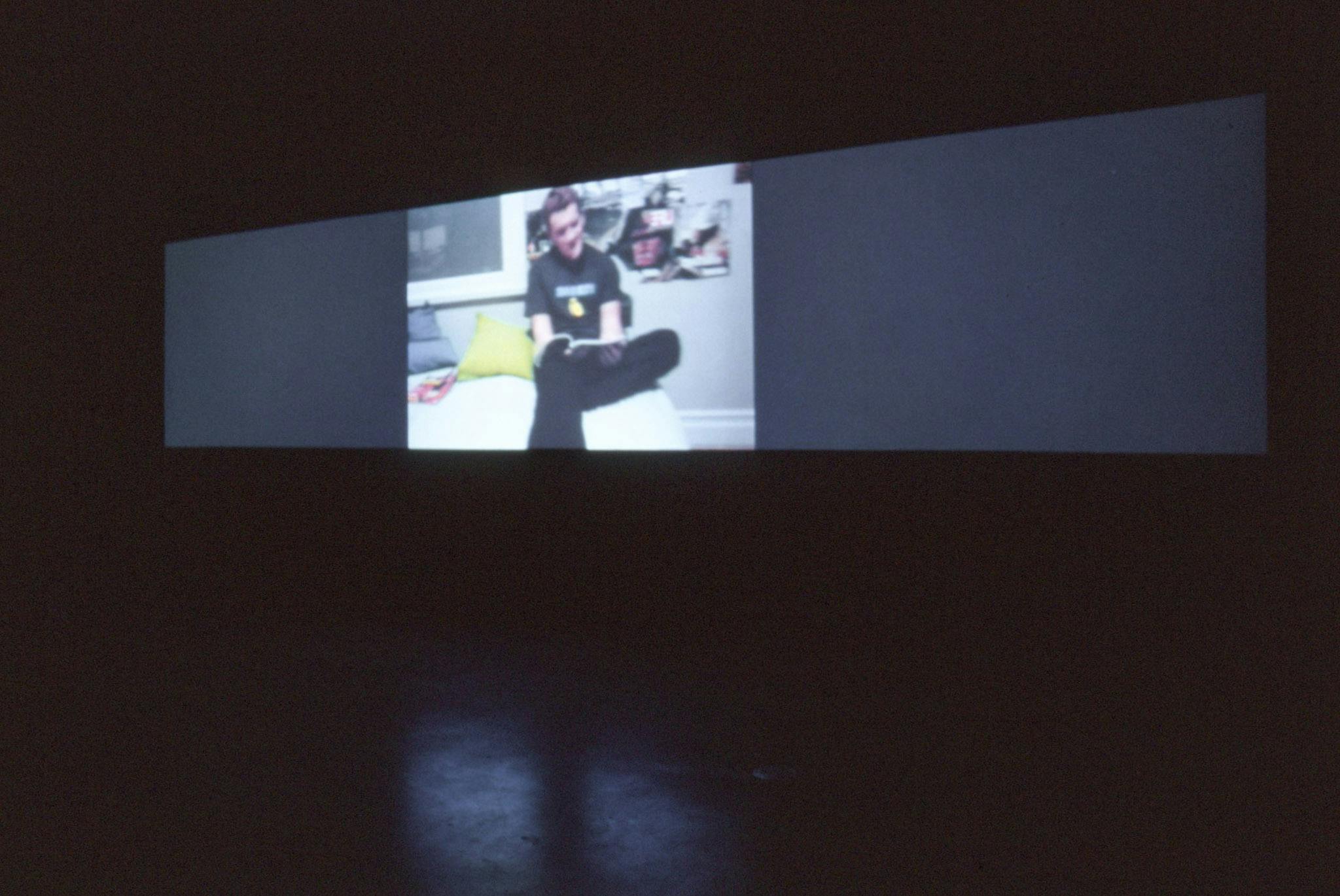 Installation image of video work by Alex Morrison. A video projected on a wall in a completely dark gallery space shows a person in a black outfit sitting at the edge of a bed reading a magazine. 