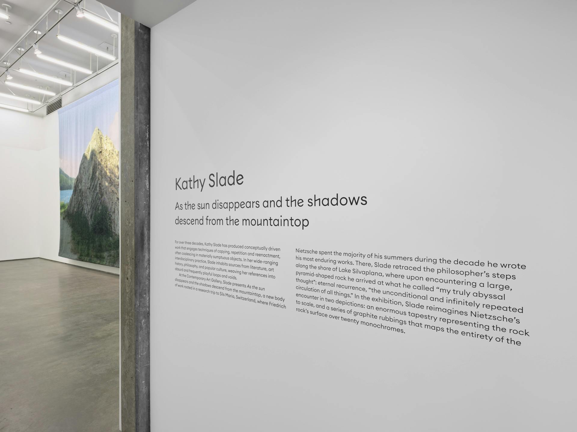 A very large tapestry by Kathy Slade depicting a large rock, visible in a gallery behind a title wall showing the exhibition's title and artist's name.