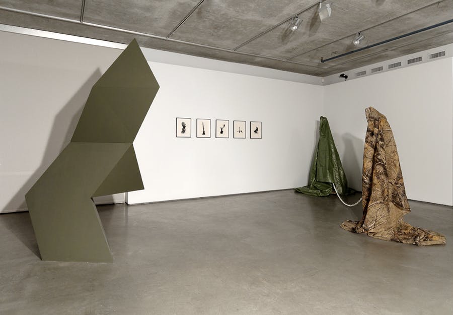 A large geometric sculpture is installed in a gallery. To the right, on the wall, five framed artworks hang. Further right, two tarp-like fabrics are draped over structures to resemble human-like forms.