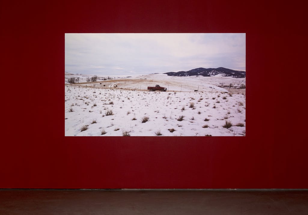 A large video projection of a snowy landscape on a red gallery wall. Centred in the image, a red pickup truck on a narrow road cutting across low, rolling hills. 