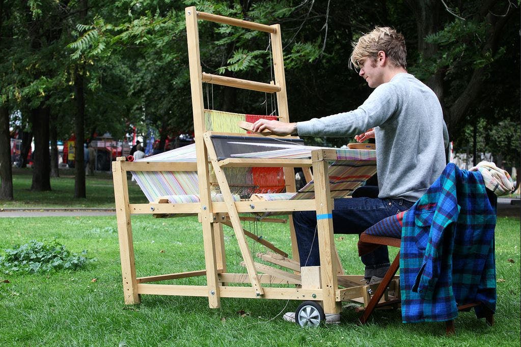 A photograph of Travis Meinolf weaving on a wooden loom outdoors.