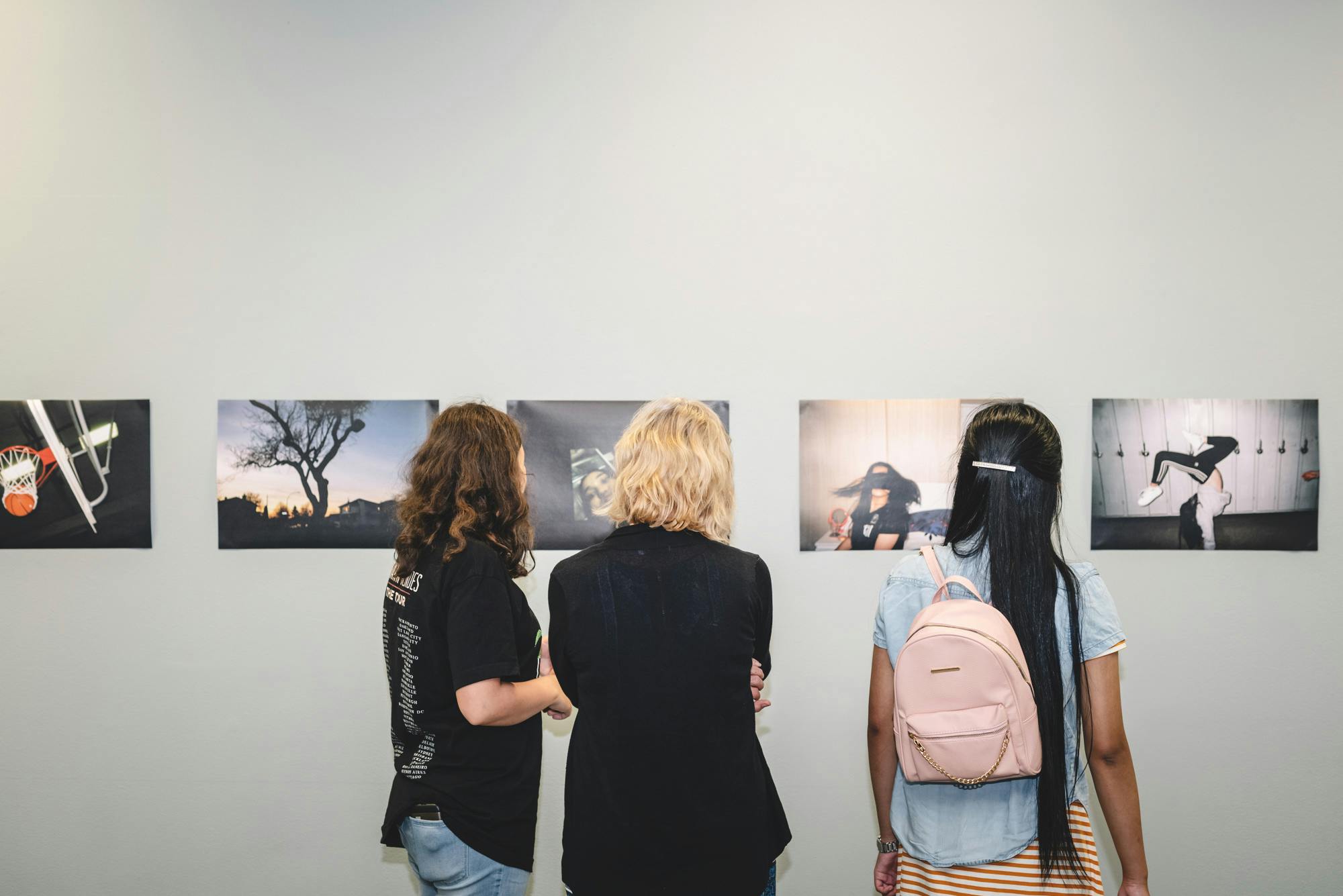 Three people, with their backs to the camera, look at a row of unframed photographs hanging on a grey gallery wall.