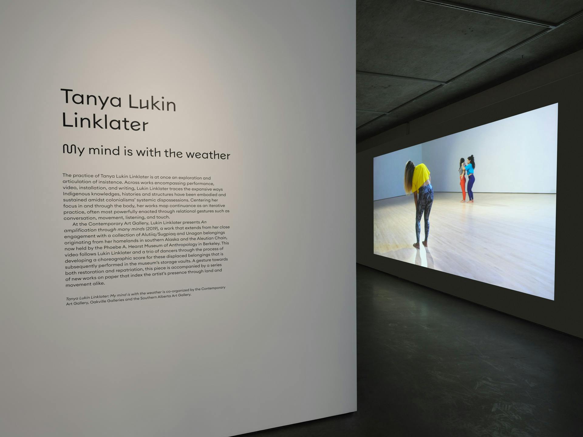 Entryway into a gallery with a video projection by Tanya Lukin Linklater visible on an interior wall. Video depicts three dancers in mid-movement in a room with white walls and wood floor.