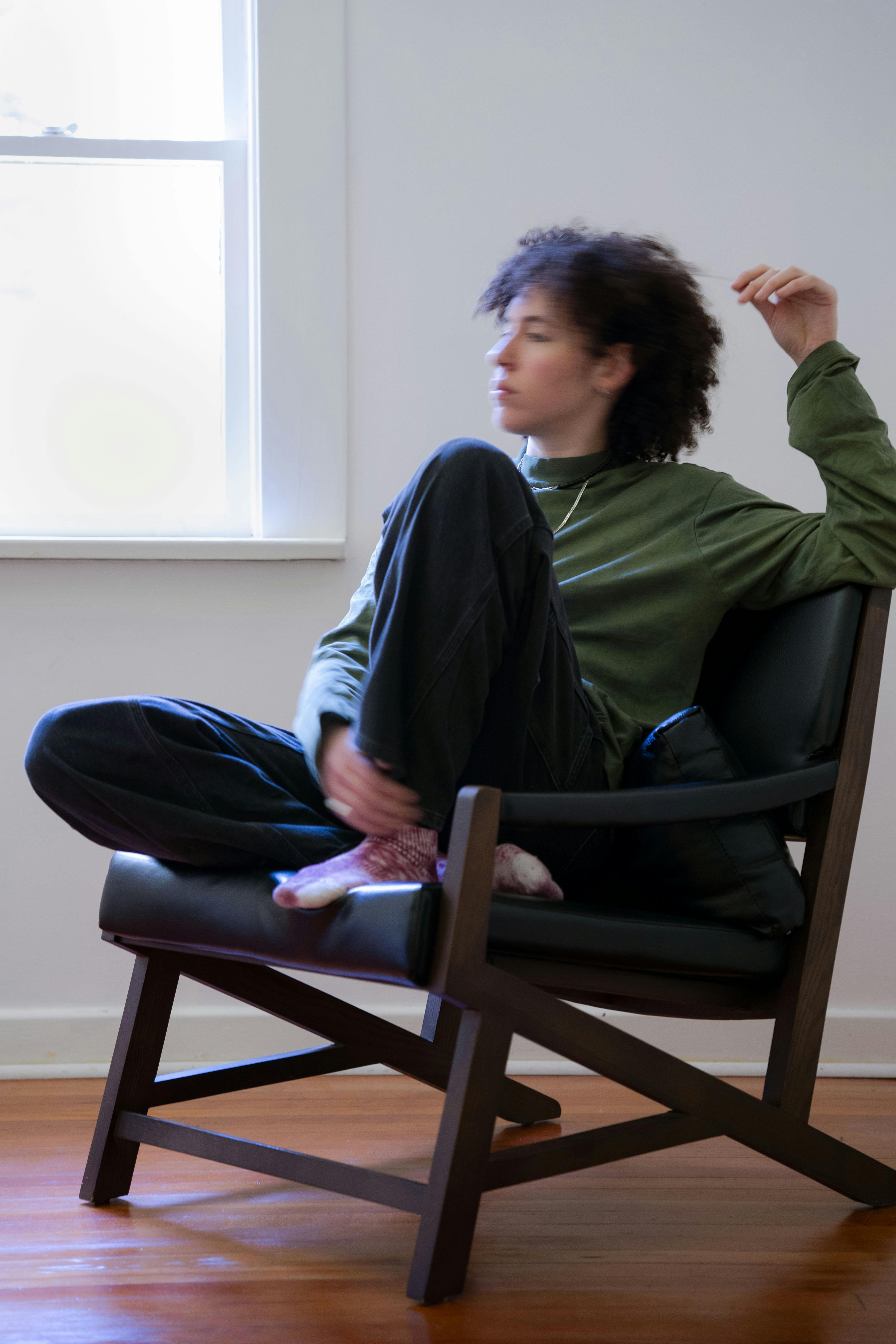 A photo of Dana Qaddah. Dana is seated in a chair wearing a green shirt and black pants. One of their arms is resting on the back of the chair.