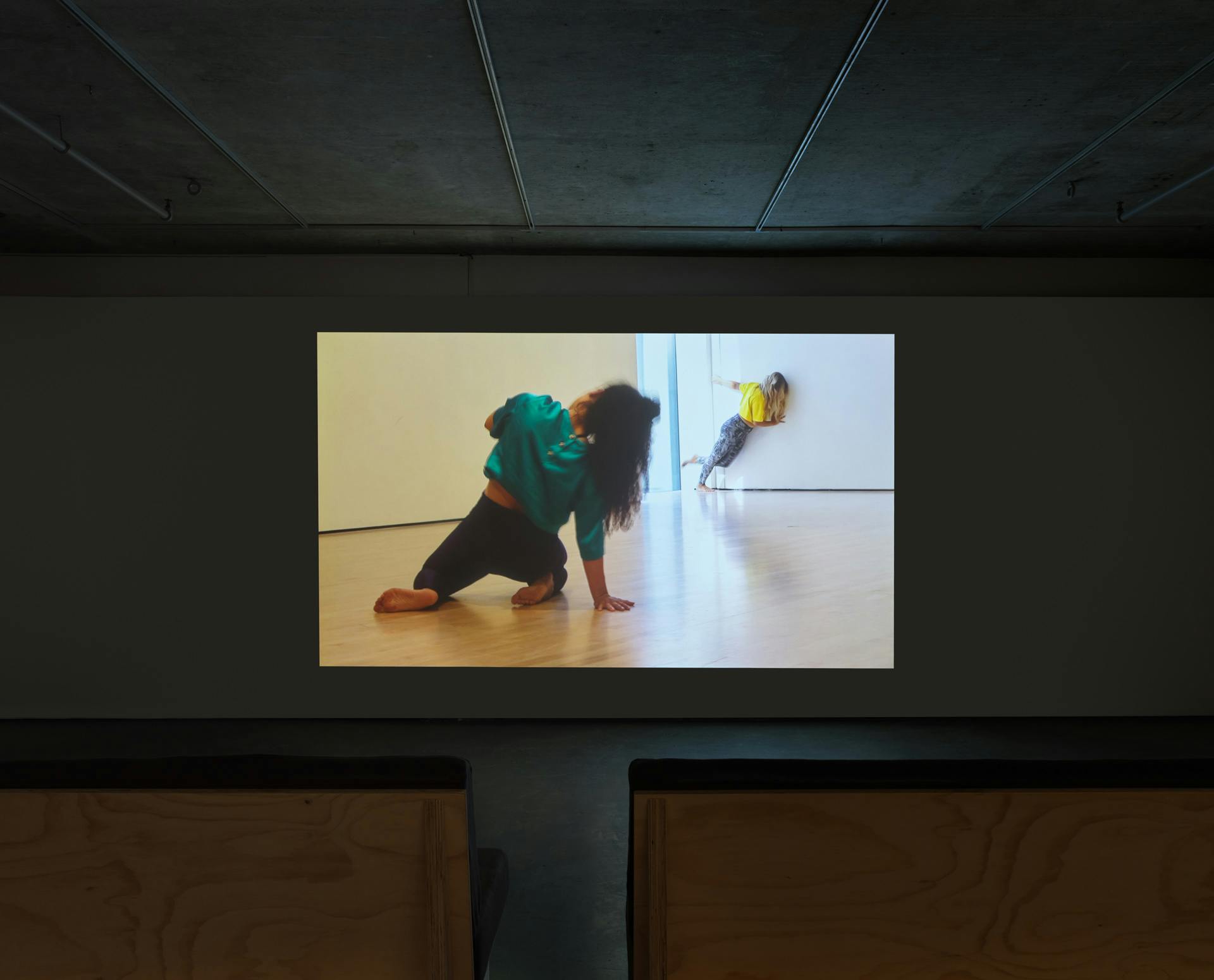 Video by Tanya Lukin Linklater projected on a wall in a dark room. Video depicts two dancers in mid-movement in a room with white walls and wood floor.