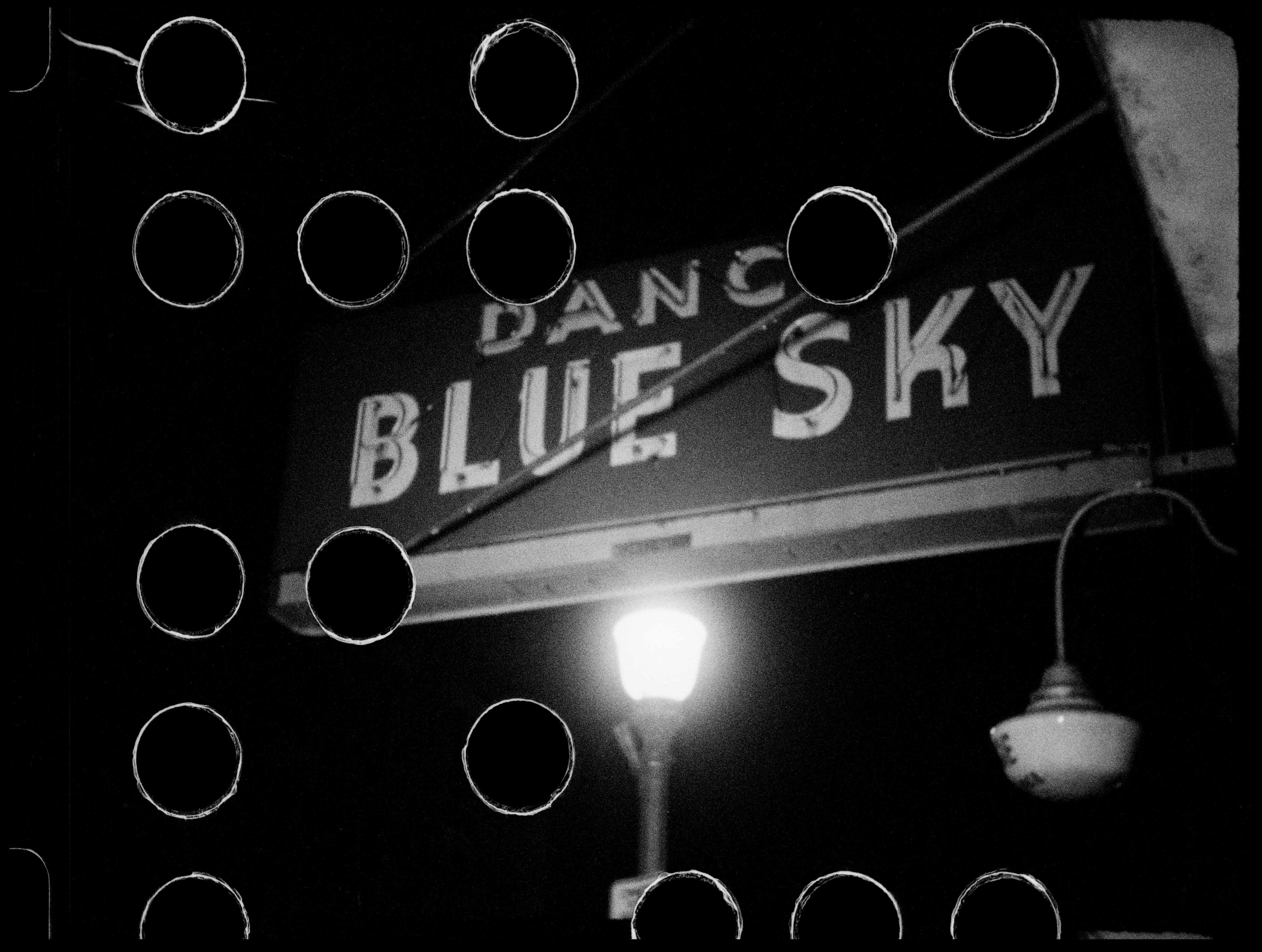 A black and white image of a film frame depicting an unlit neon sign and street light at night. The neon sign reads “DANC BLUE SKY.” Black circles cover the surface of the image. 