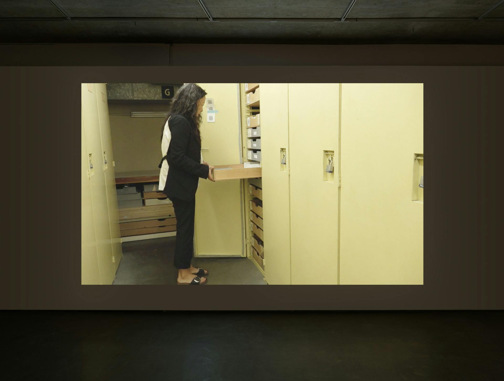 Video by Tanya Lukin Linklater projected on a wall in a dark room. Video depicts a woman opening a drawer inside an open storage locker surrounded by two rows of closed storage lockers.