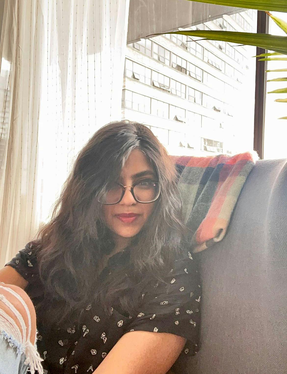 A photo of Aakansha Ghosh. She is seated on a gray couch wearing a black patterned top and dark rimmed glasses.