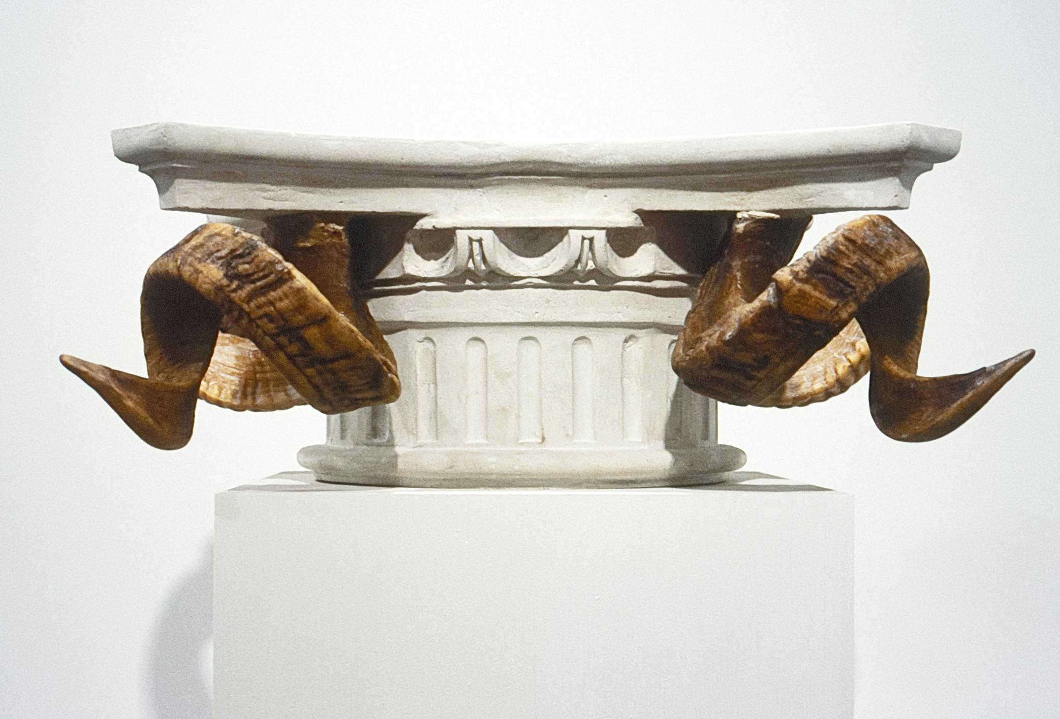 A close up view of the top of a capital column sculpture. Ram horns protrude from the sides of the column. 