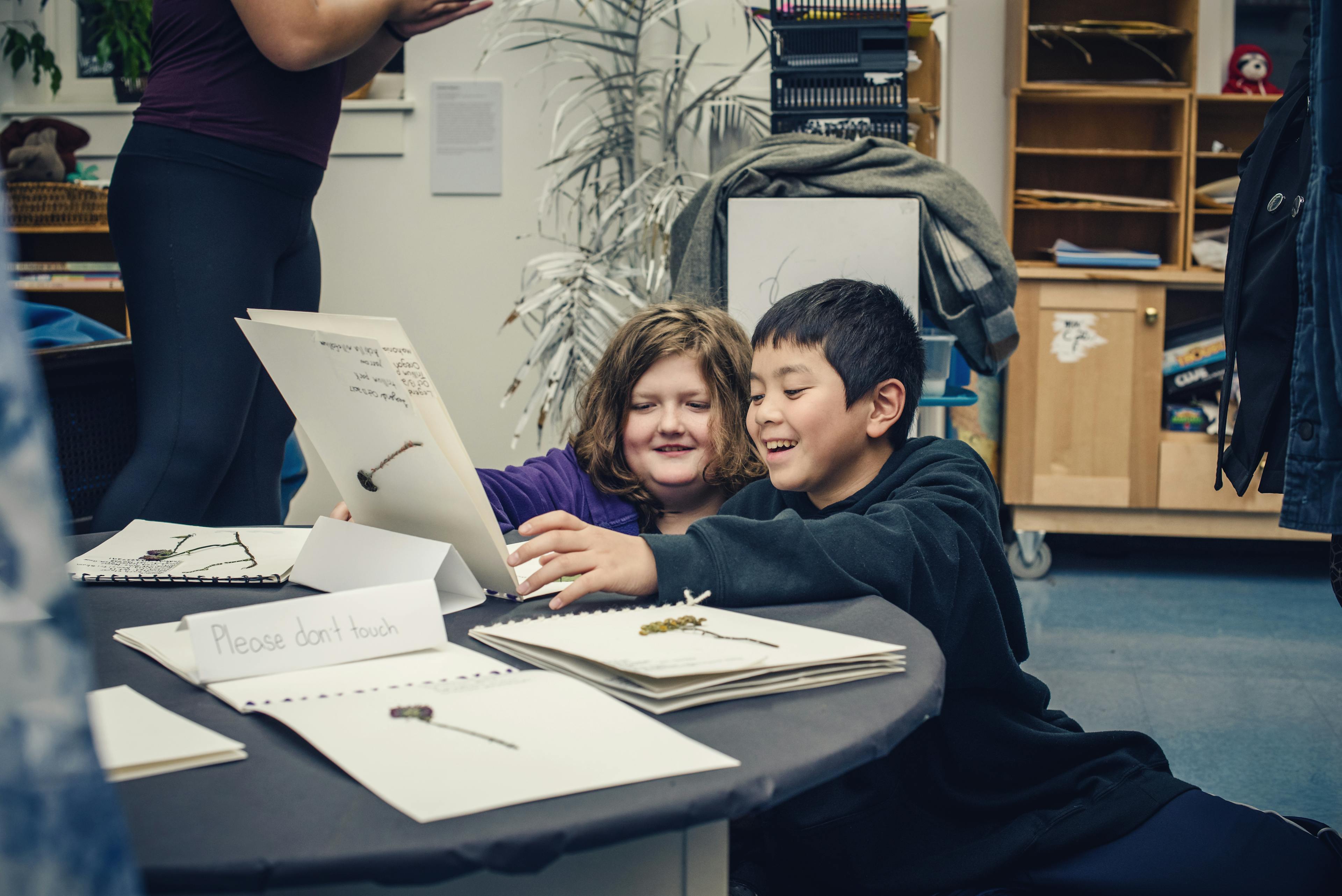 Two smiling children in a classroom looking at a handmade herbarium book. Other herbarium books surround them on the table.   